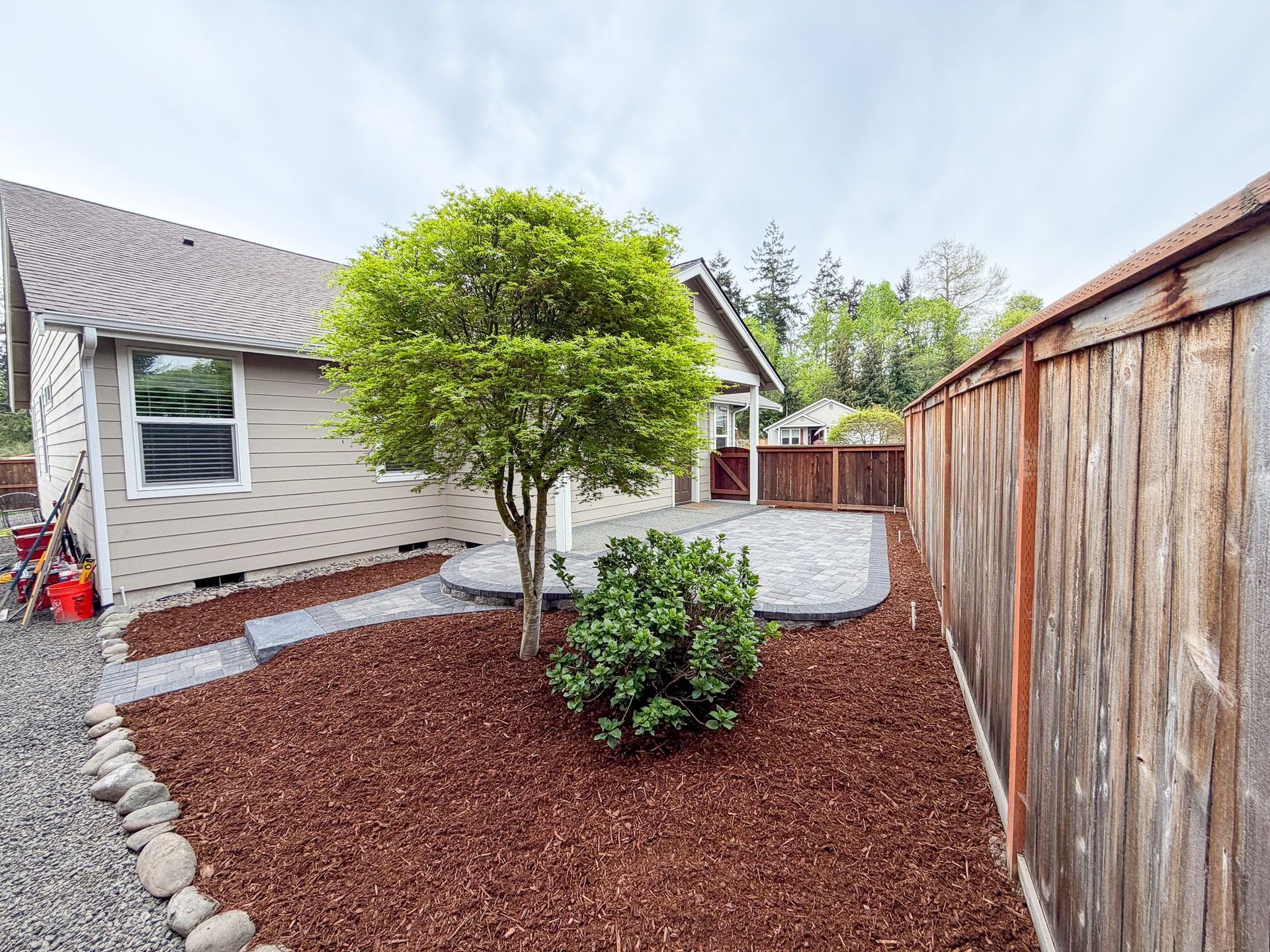Landscaped backyard with a tree, shrubs, stone border, and a wooden fence beside a house