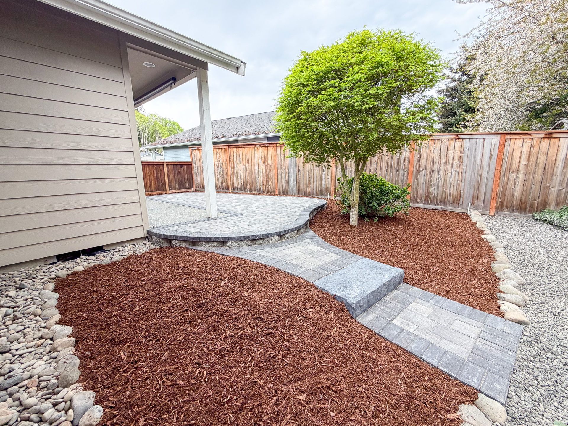 Landscaped backyard with red mulch, stepping stones, a small tree, and a wooden fence beside a house