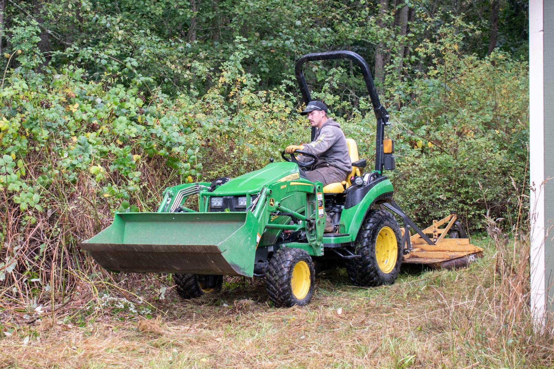 Man operating a green John Deere tractor with a front loader and rear mower, clearing brush.