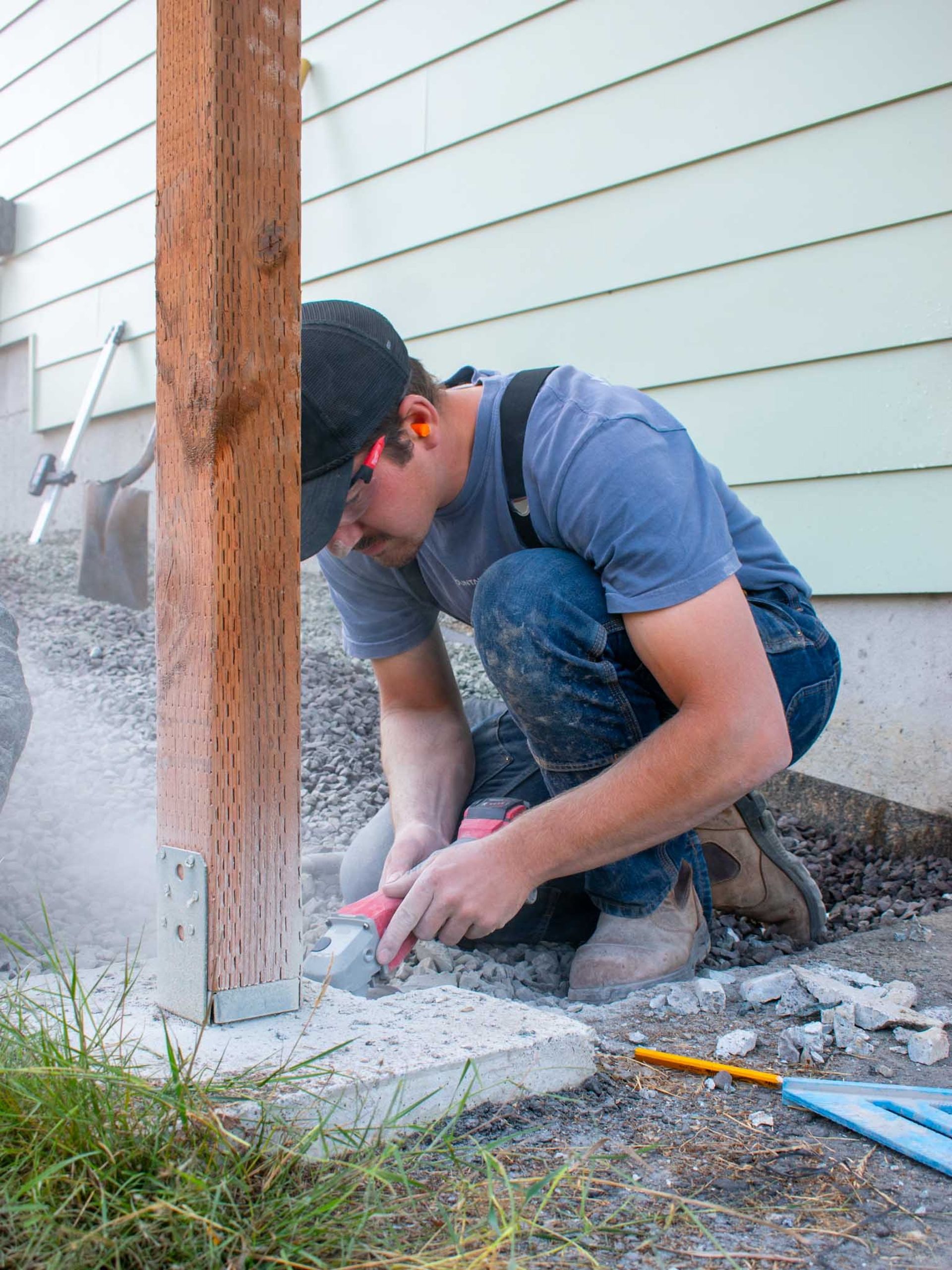 Person using a power tool to cut concrete at the base of a wooden post outdoors.