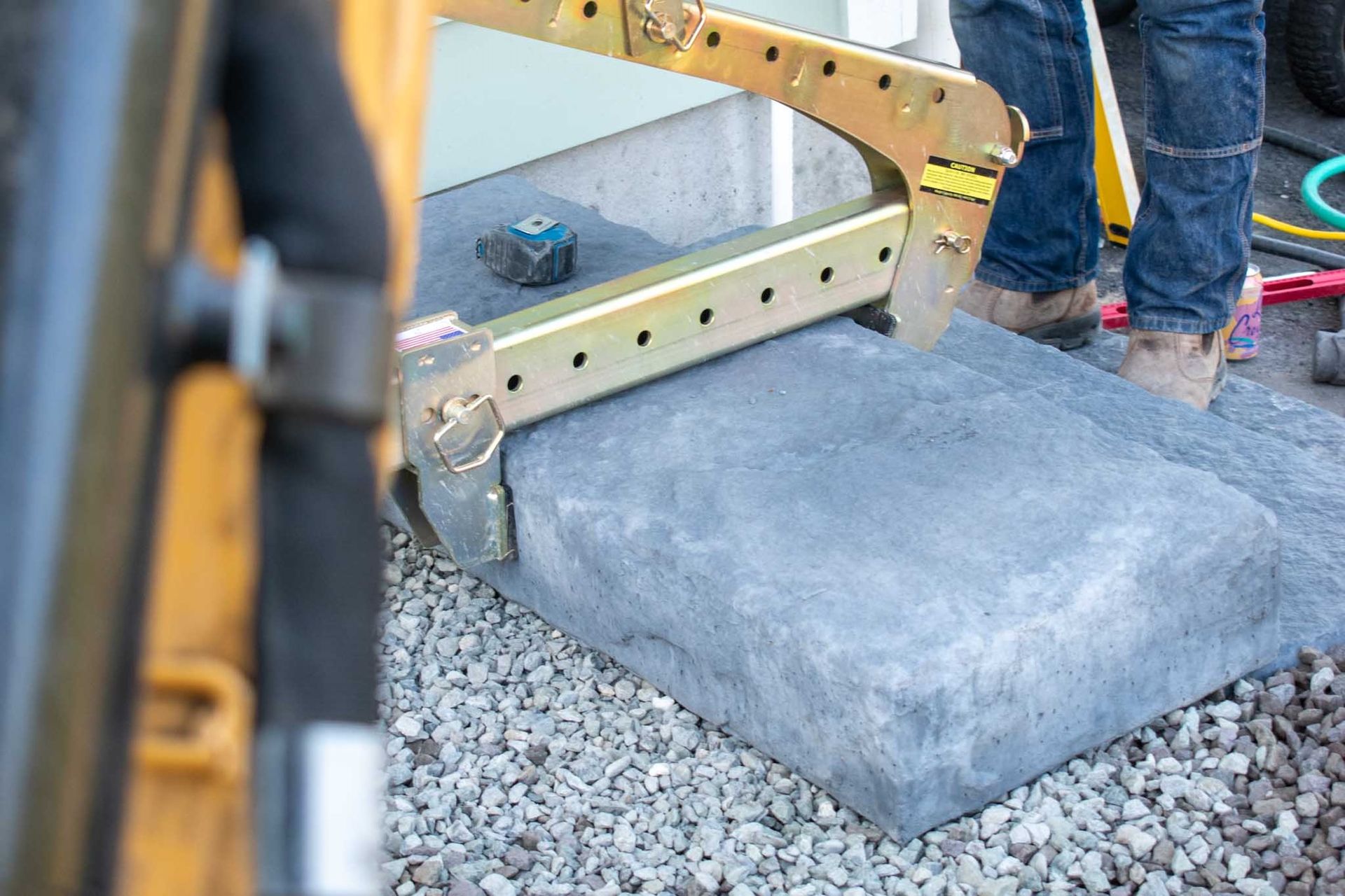 Worker using a clamp to lift a large gray stone paver; construction site setting.