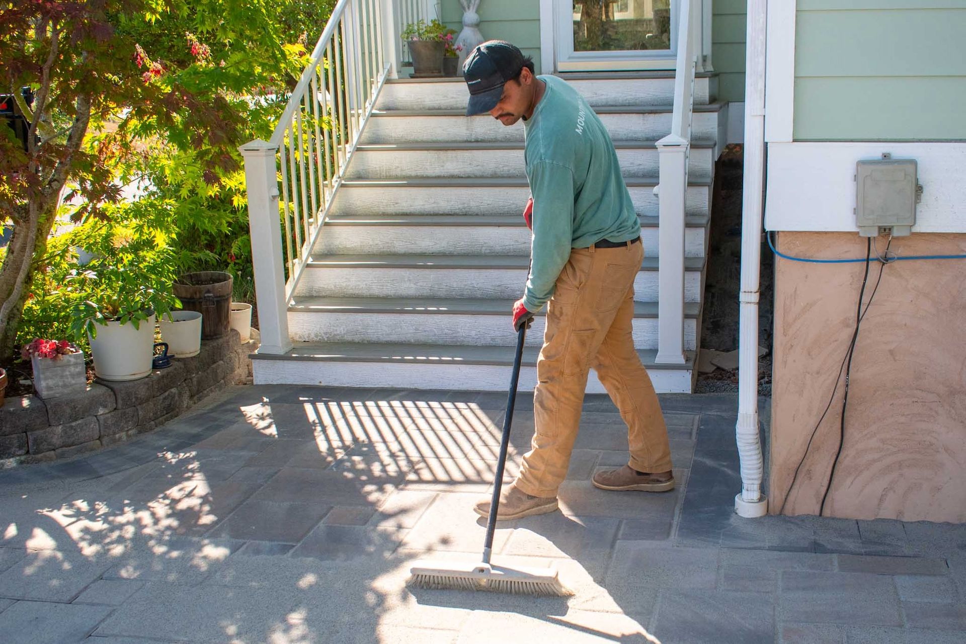A person sweeps a stone patio near white stairs, wearing a long-sleeved shirt and hat.