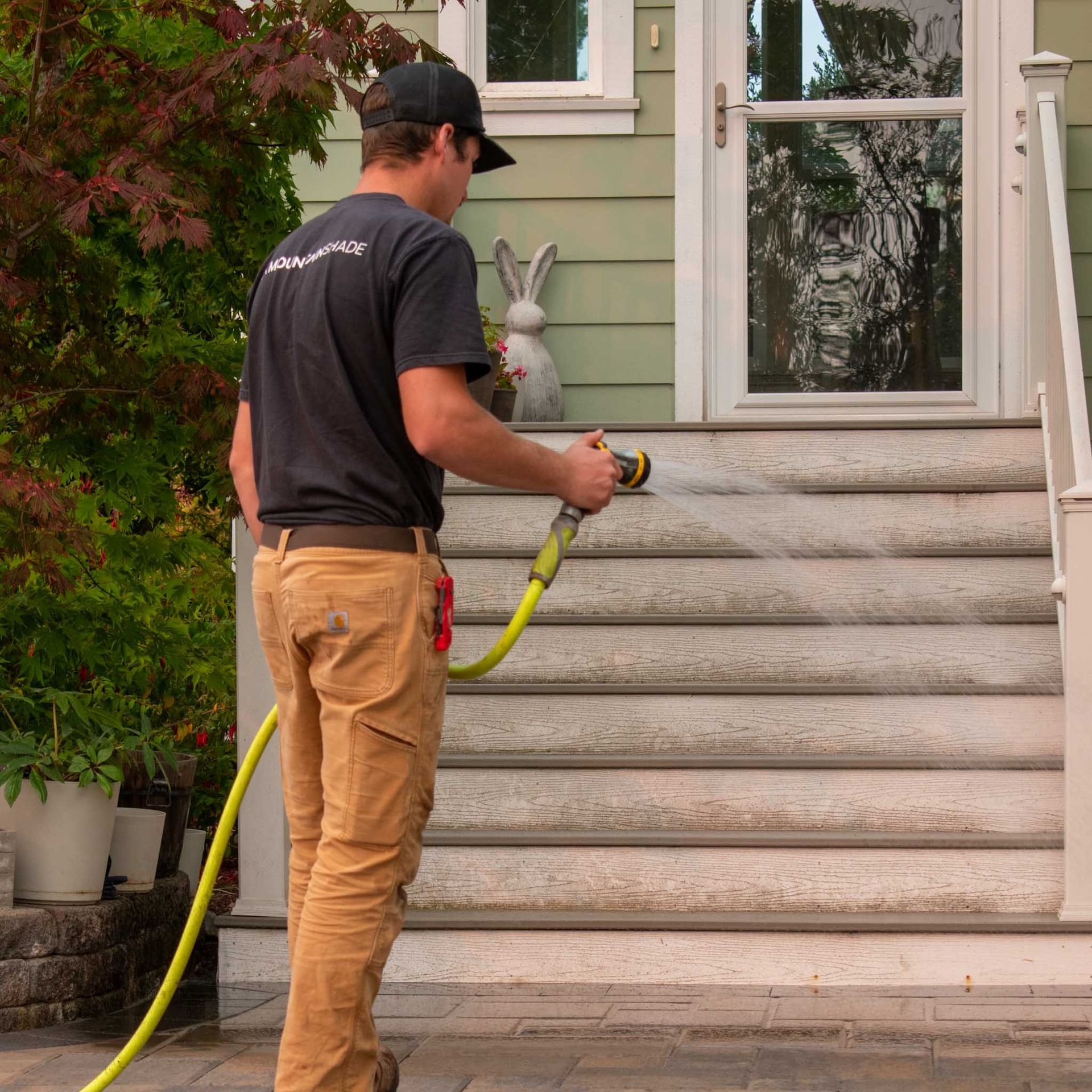 Man in work clothes sprays water on outdoor steps with a hose.