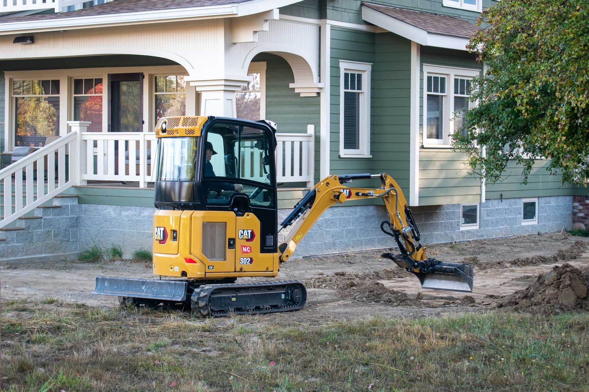 Yellow excavator digging dirt near a green house with white trim.