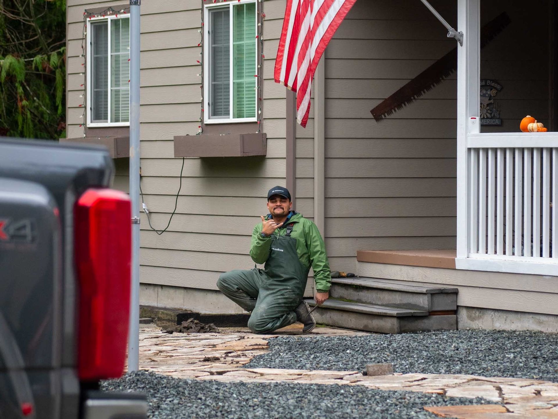 Man in green overalls kneels, holding a device near a house with an American flag.