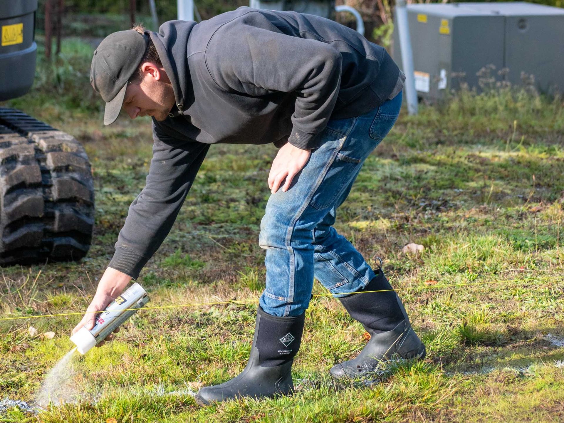 Man in a black hoodie and boots pouring a substance from a container onto grass near construction equipment.