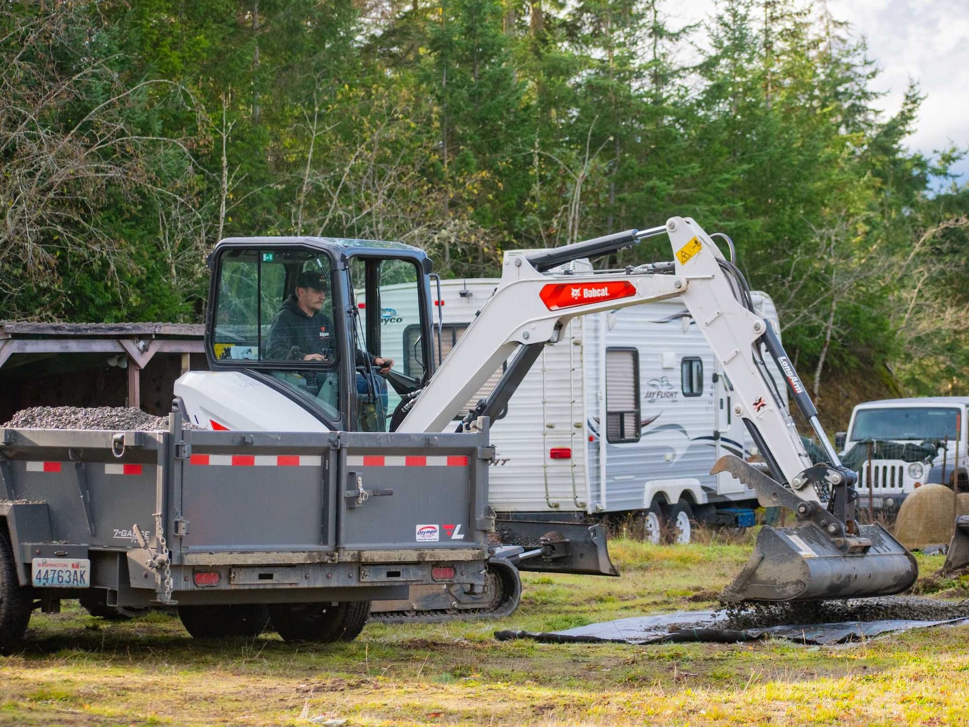 White excavator on trailer, operator inside. Dirt and trailers in background.
