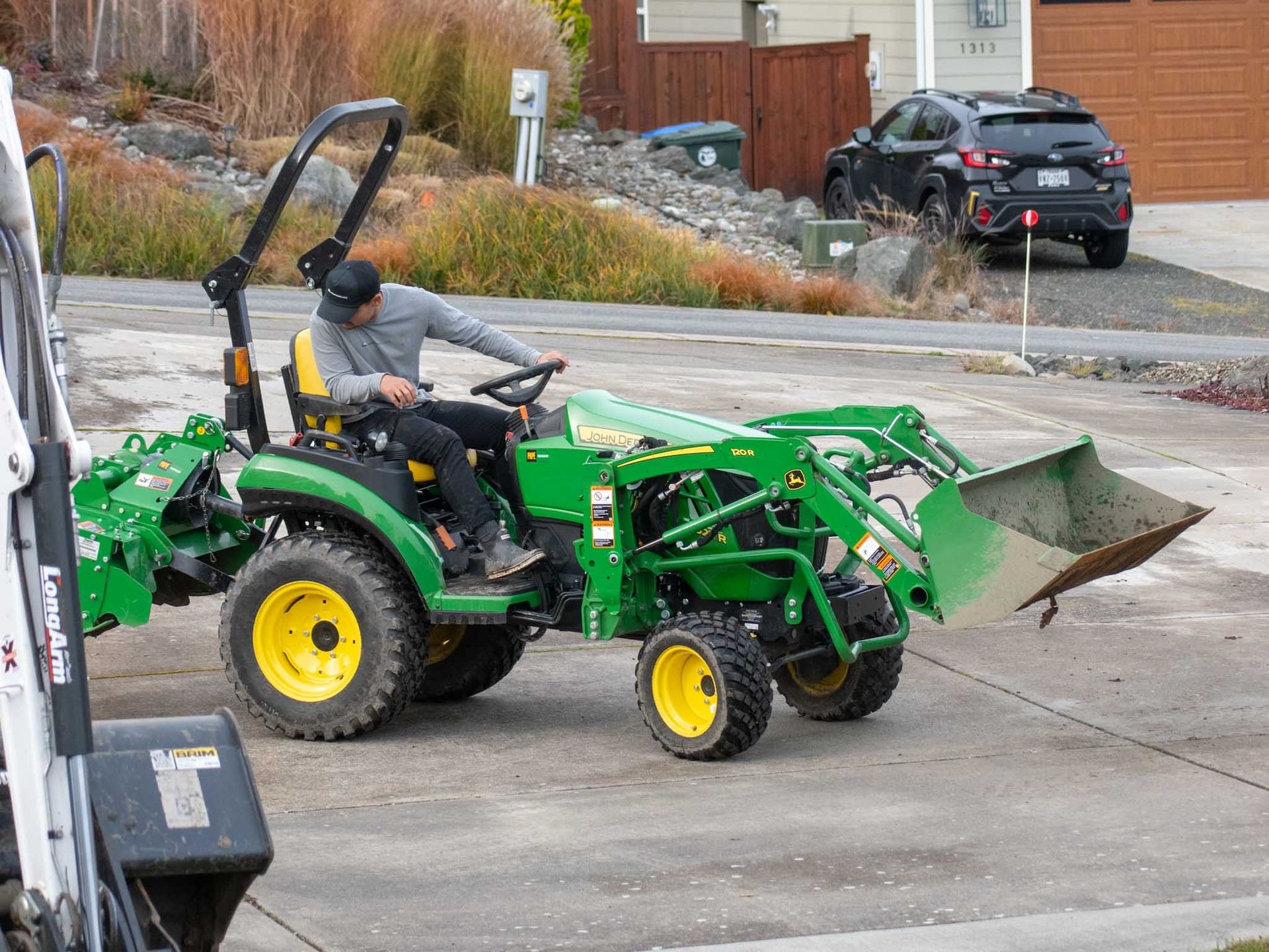 Person operating a green John Deere tractor with a front loader and rear attachment outdoors.