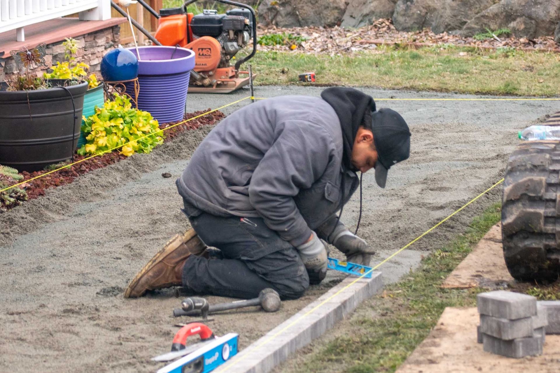 Person kneels, using tools to install pavers on a gravel bed; outdoor setting with machinery and plants.