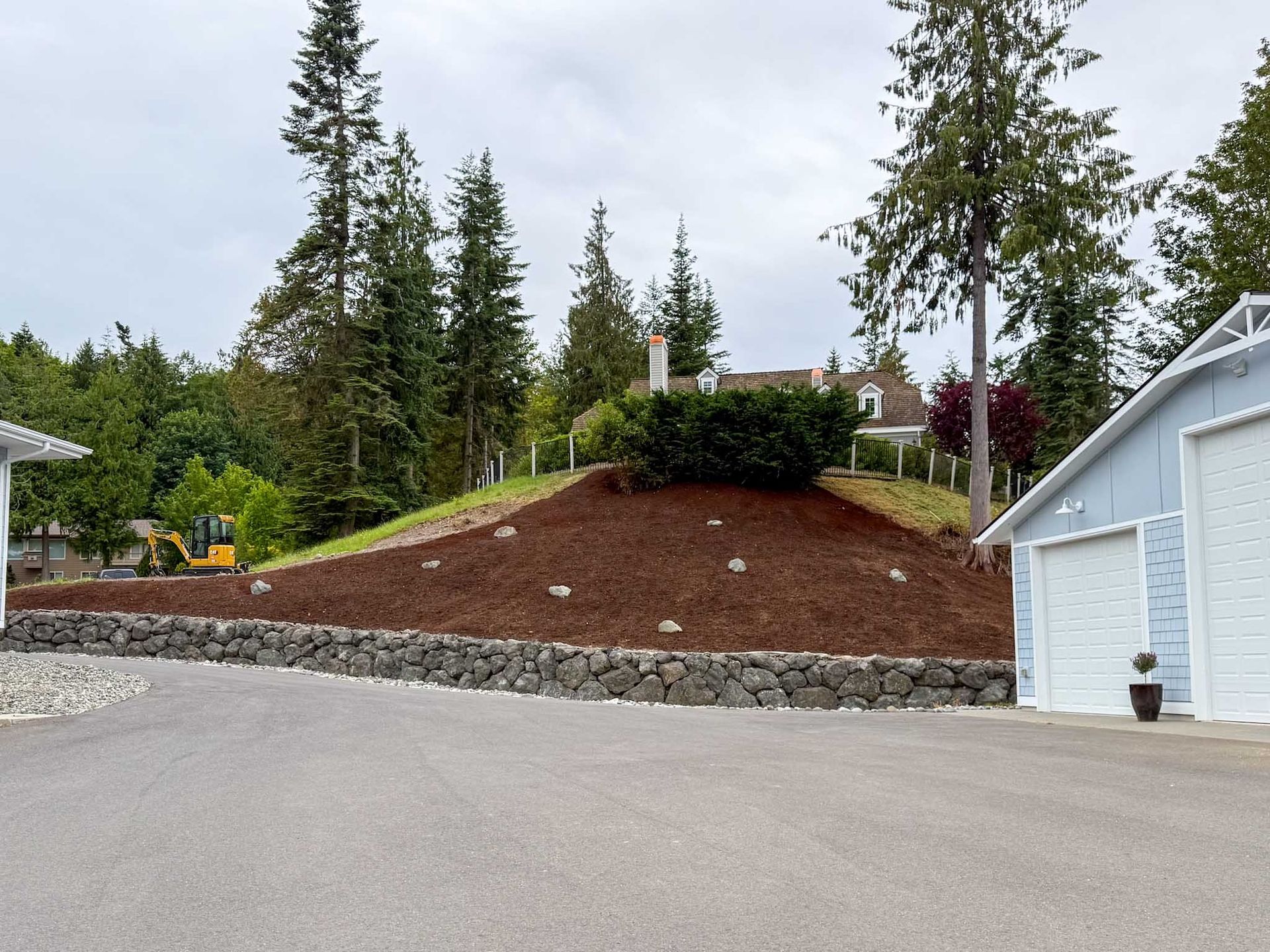 Asphalt driveway with retaining wall, mulched hill, light blue garage, and trees in background.