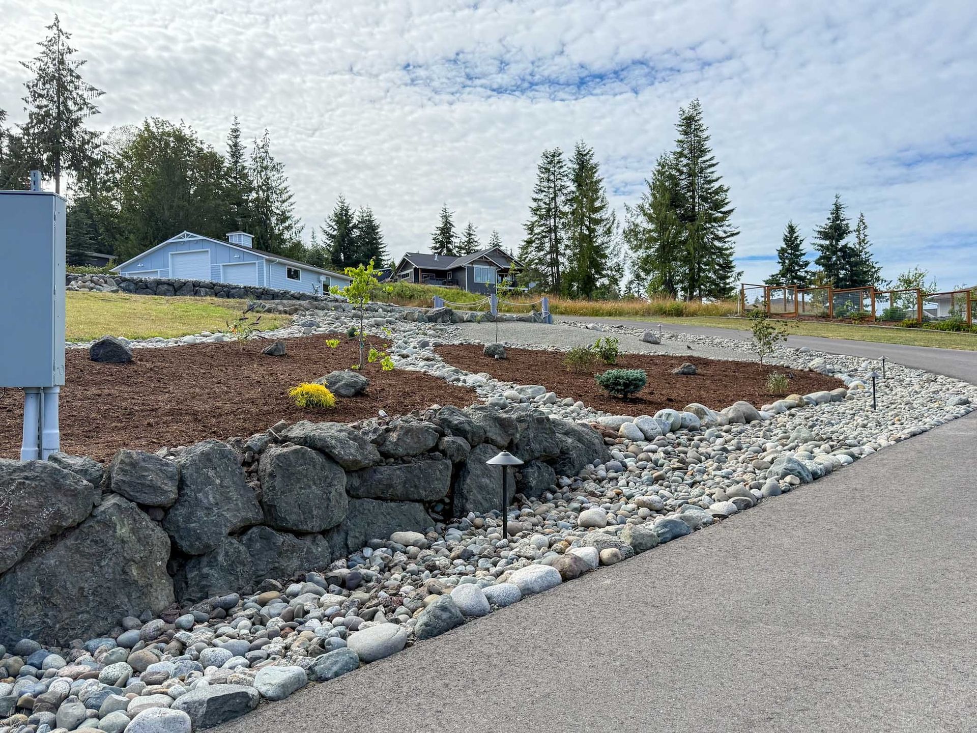 Stone wall and gravel border a driveway lined with mulch and small plants, a house sits atop a hill.