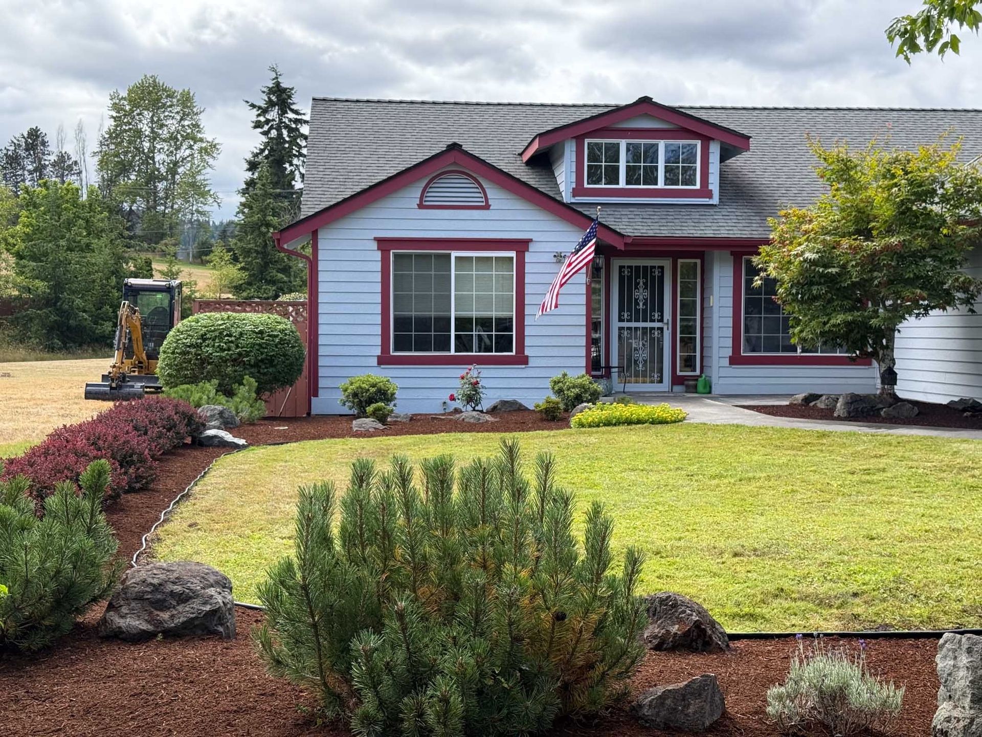 Blue house with red trim, American flag, green lawn, landscaping with small excavator in background.