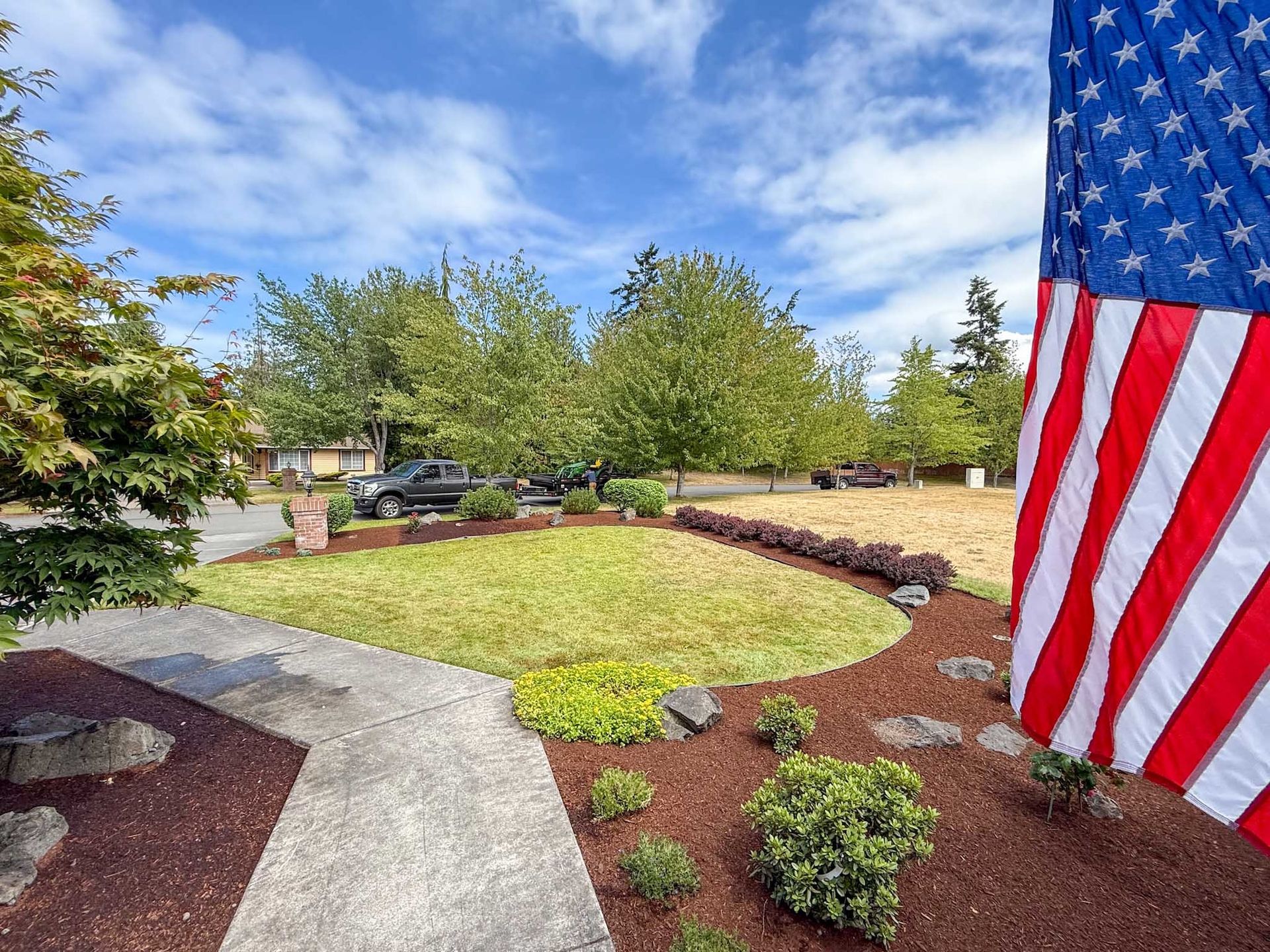 Lush lawn with flower bed and mulch, American flag in the right corner, blue sky with some clouds, residential setting.