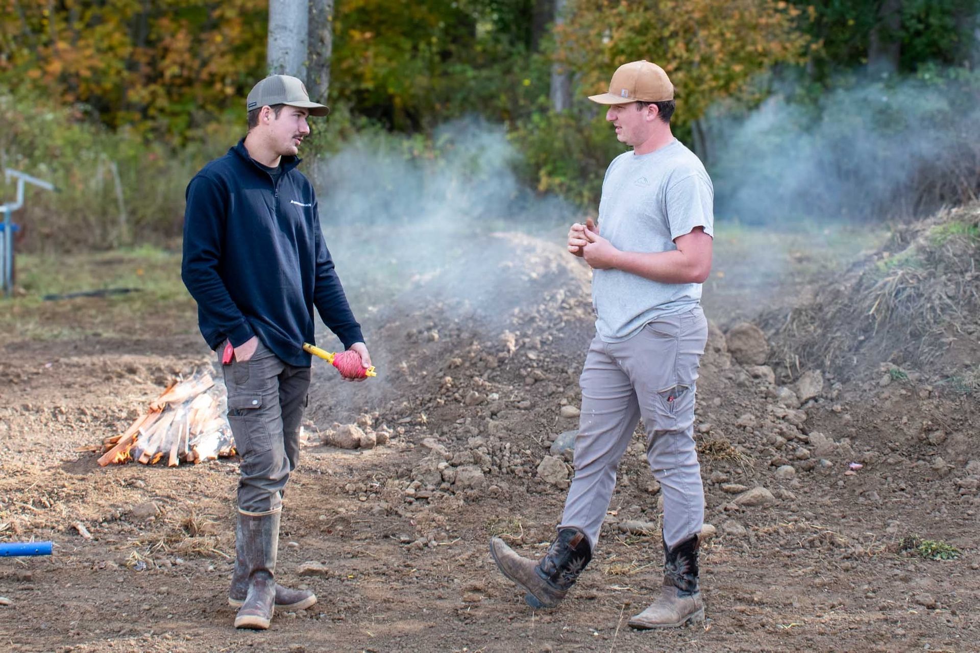 Two men stand talking outside, next to a small fire and dirt pile. One holds a tool.