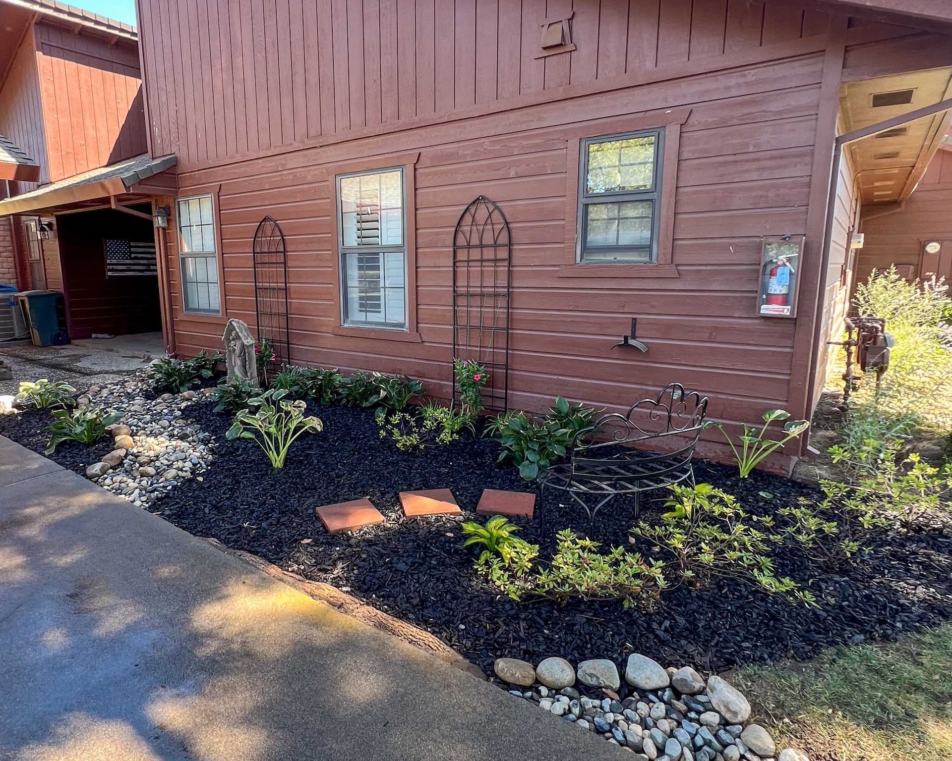 Exterior of a reddish-brown building with a landscaped garden bed filled with plants, mulch, and stepping stones.