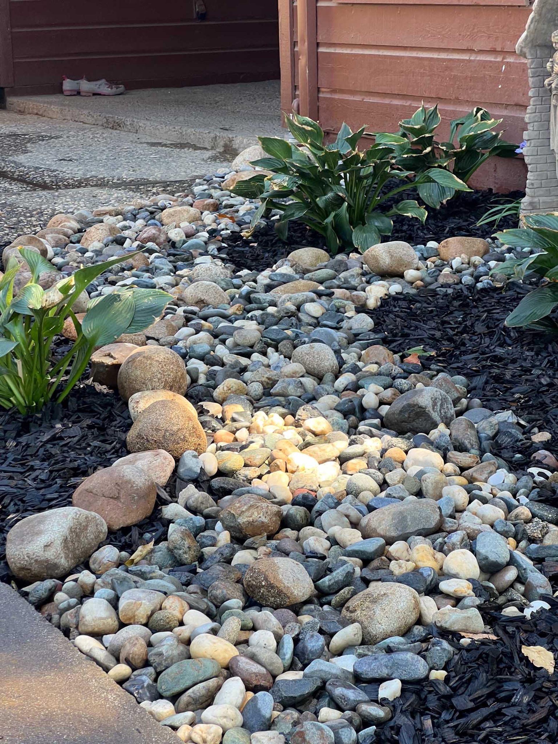 A decorative garden bed with a dry creek bed of rocks surrounded by mulch and greenery.
