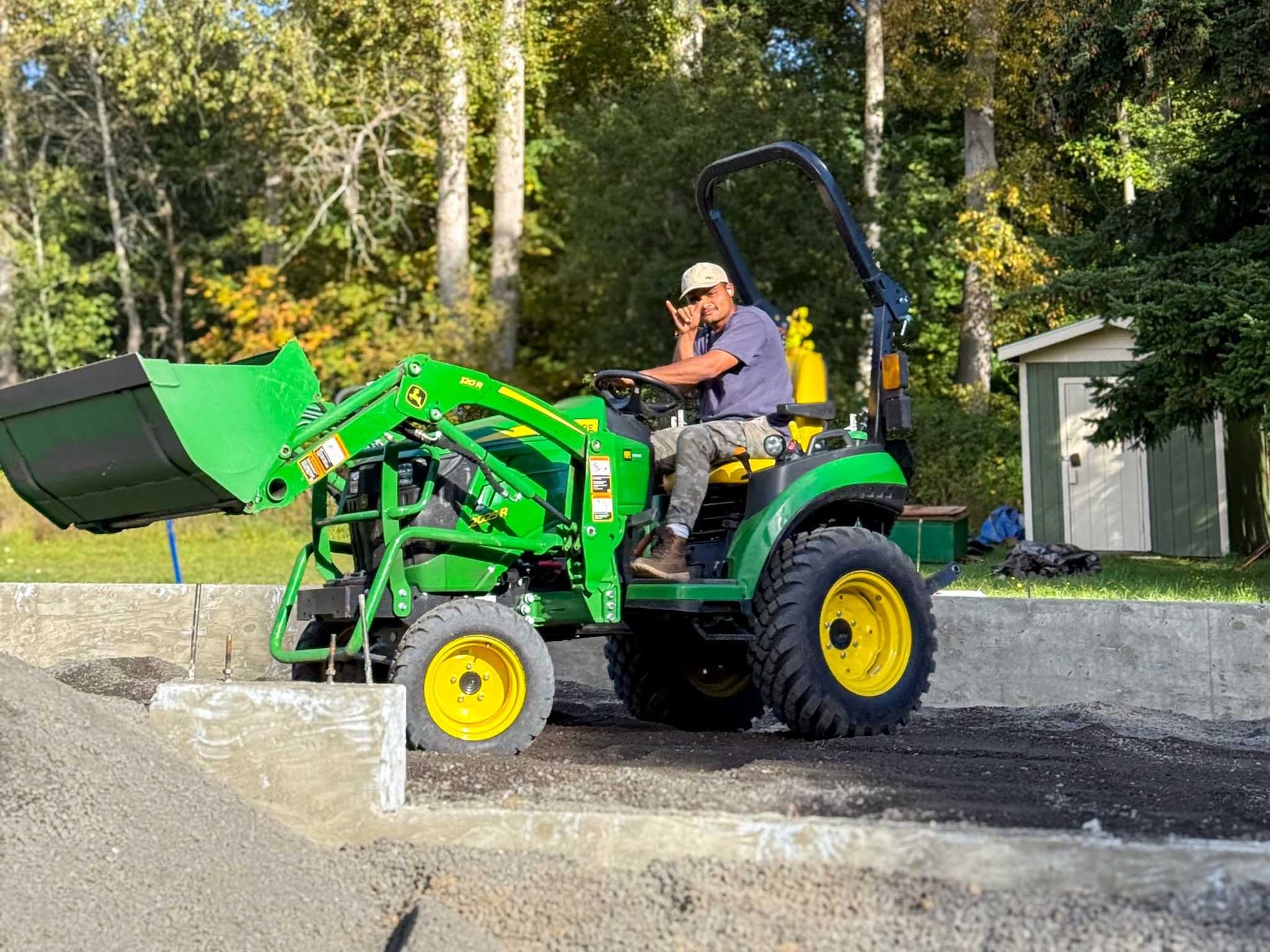 Man operating a green John Deere tractor with a front loader. He’s outside near a pile of gravel and a small shed.