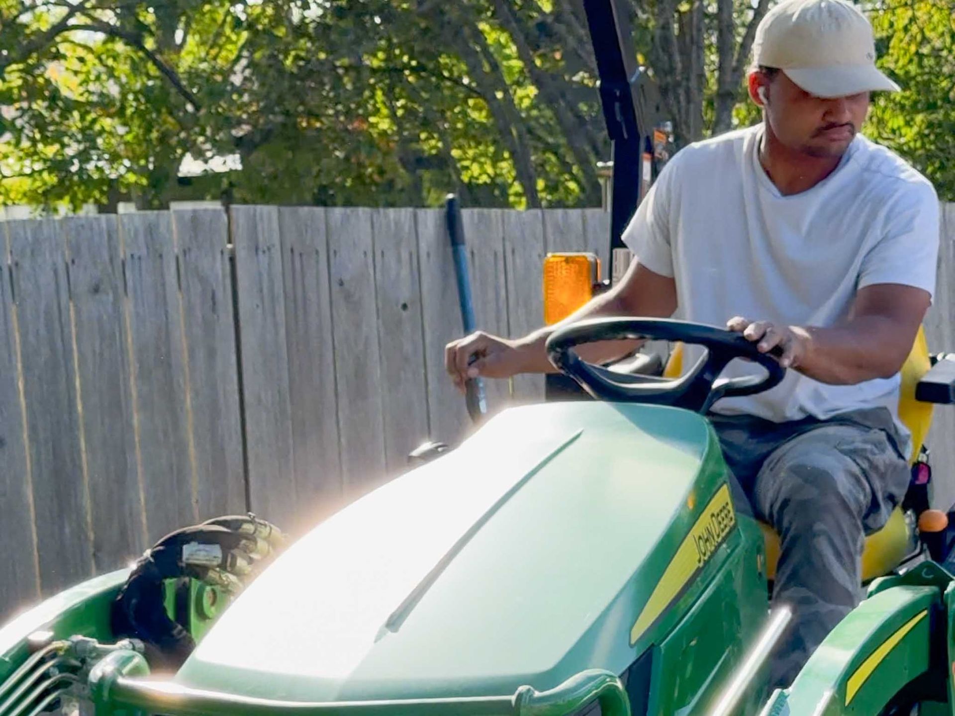 Man driving a green tractor with a beige cap near a wooden fence.