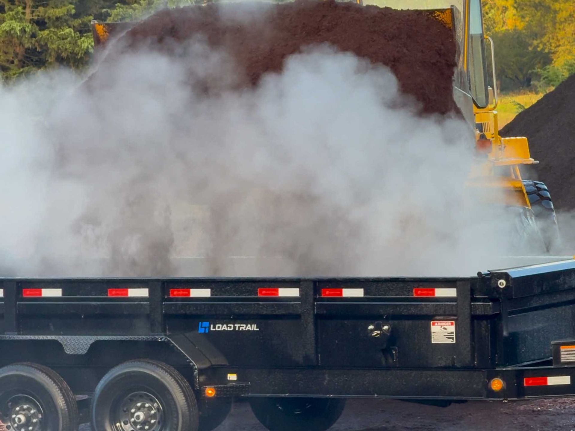 A front-end loader dumping steaming mulch into a black trailer.