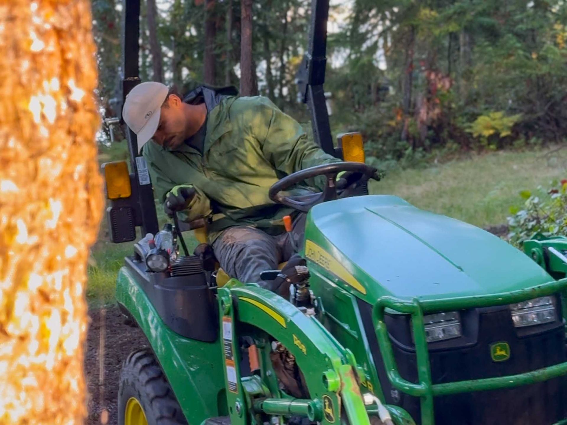 Man on a green John Deere tractor in a wooded area, operating controls, wearing a jacket and cap.