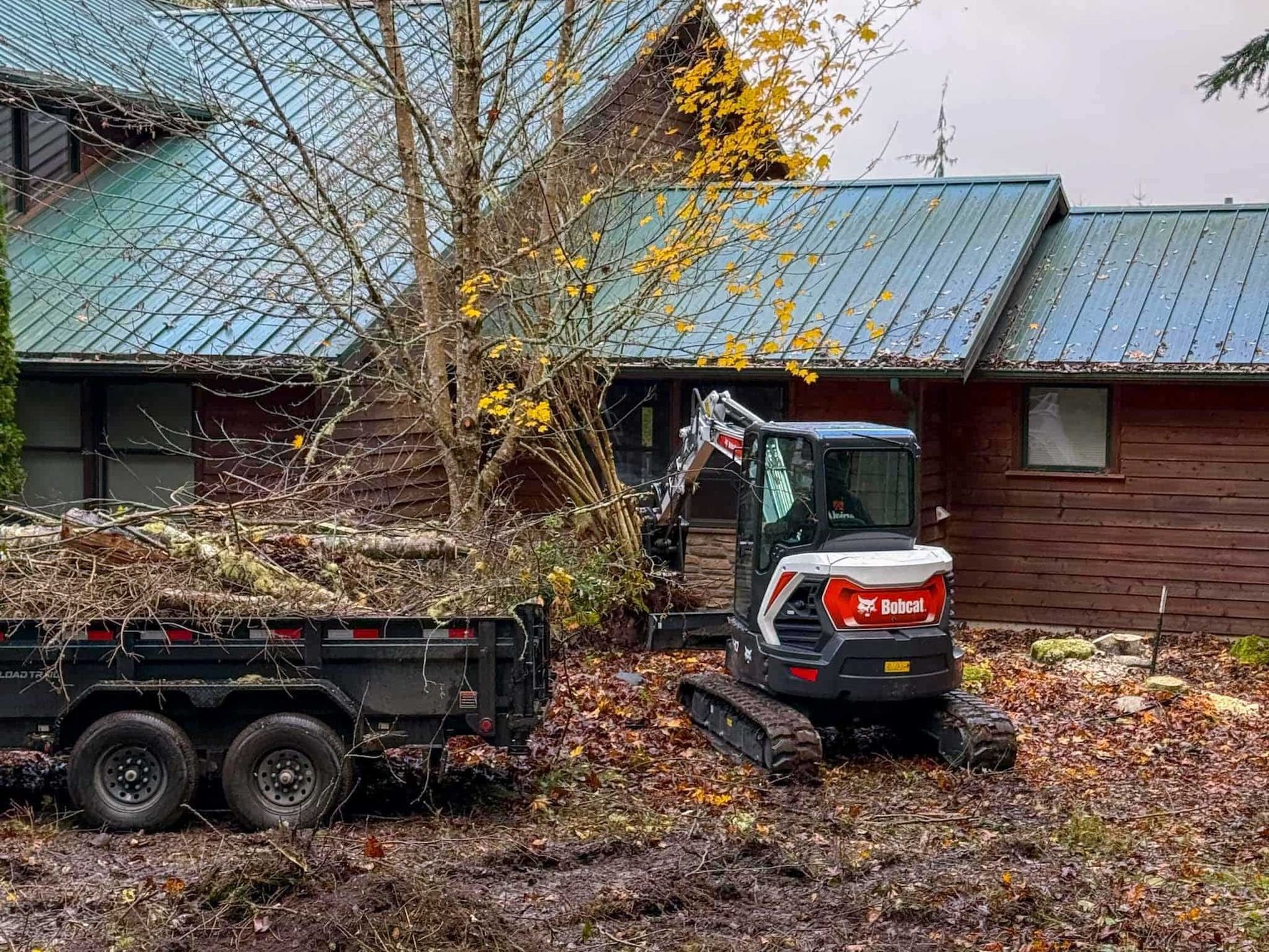Bobcat excavator and trailer beside a wooden house, clearing brush and debris.
