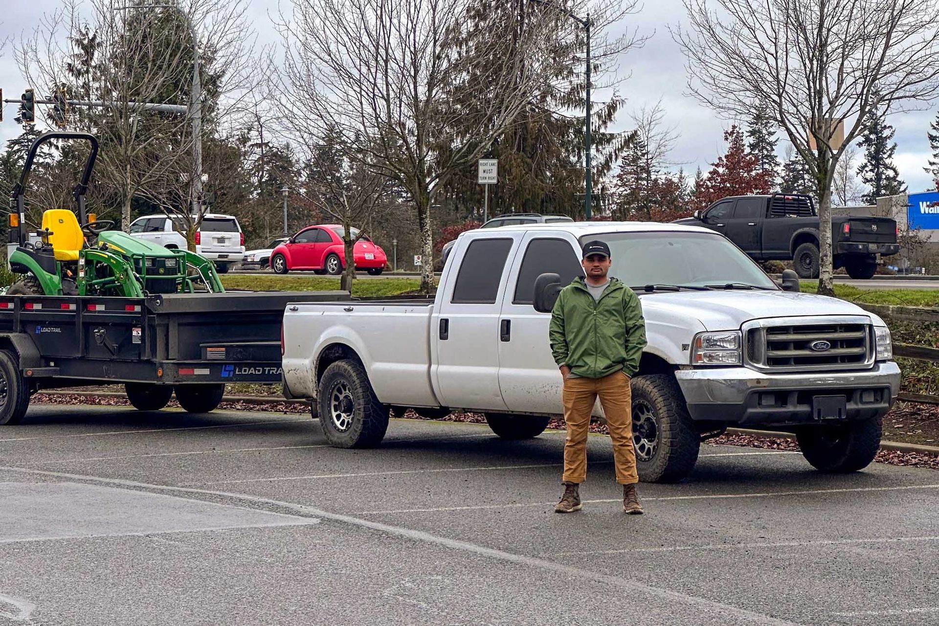 Man standing in front of a white truck towing a trailer with a green tractor.