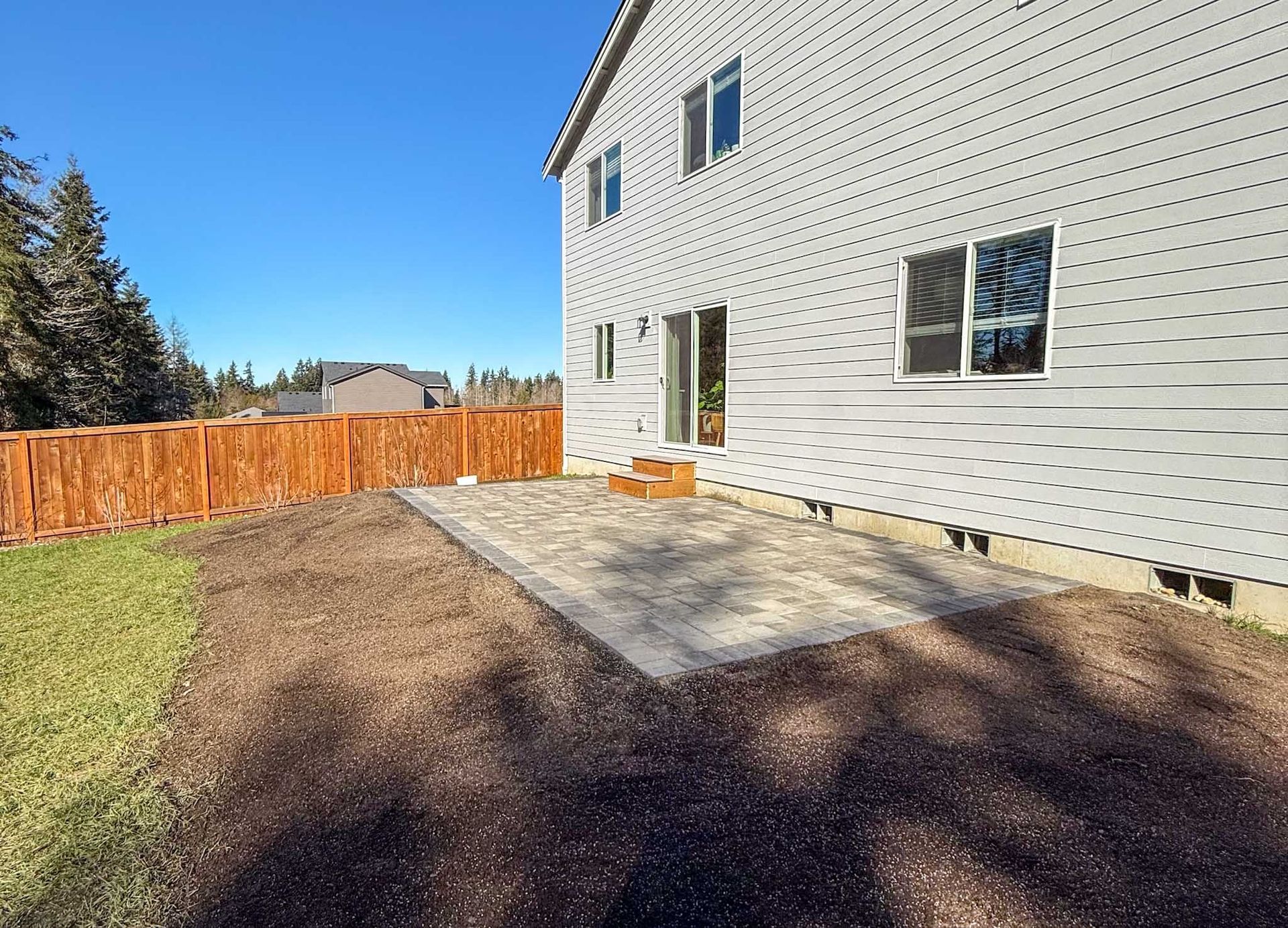 Backyard with a concrete patio, wooden fence, and light gray house on a sunny day.