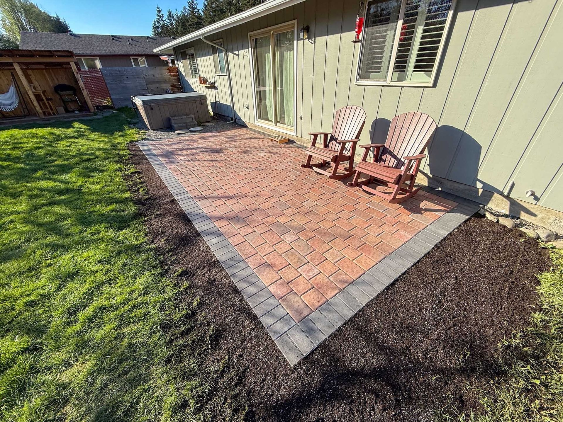 Brick patio with two wooden chairs next to a house with a sliding glass door and bark mulch border.