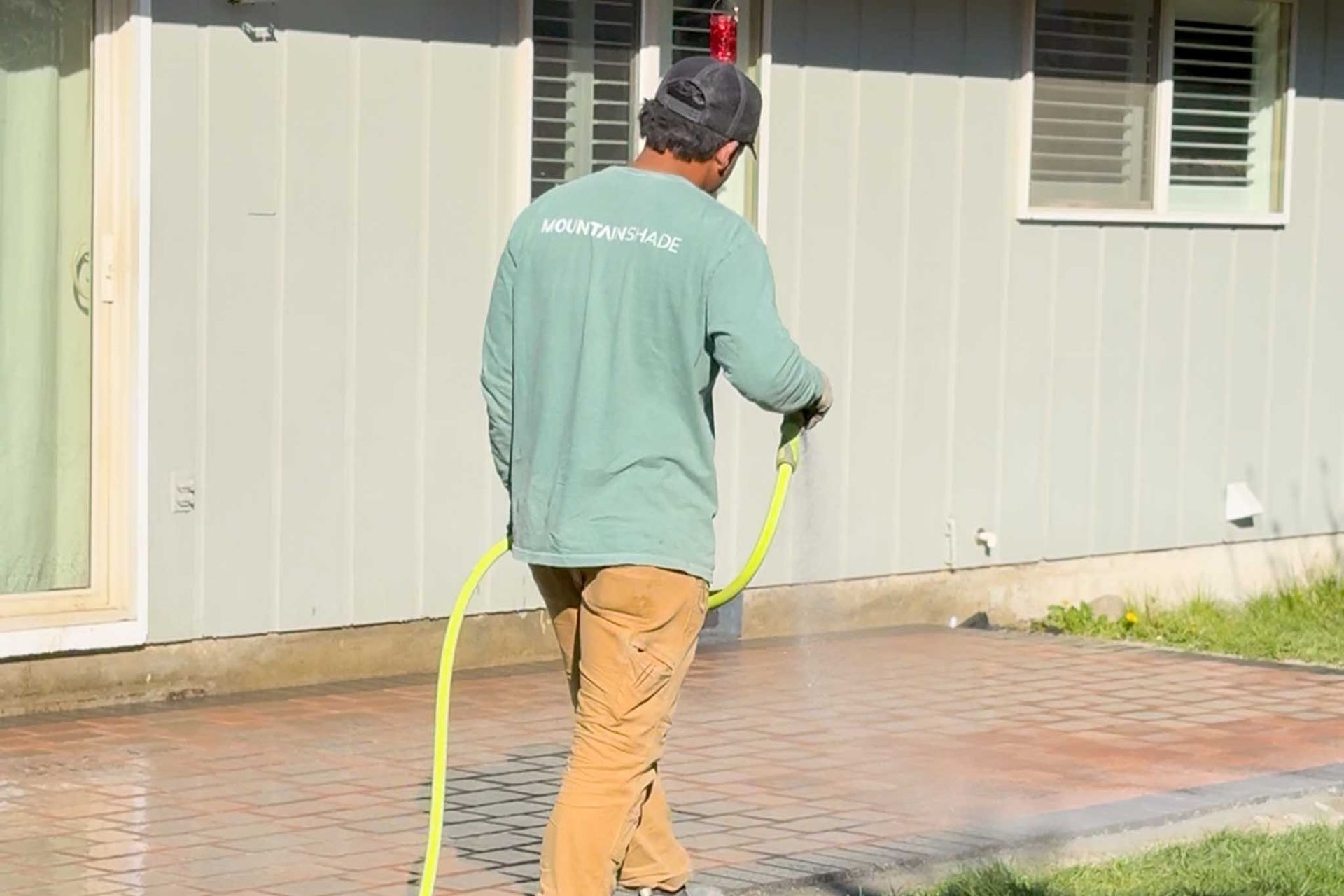Man in green shirt hoses down a brick patio next to a house.