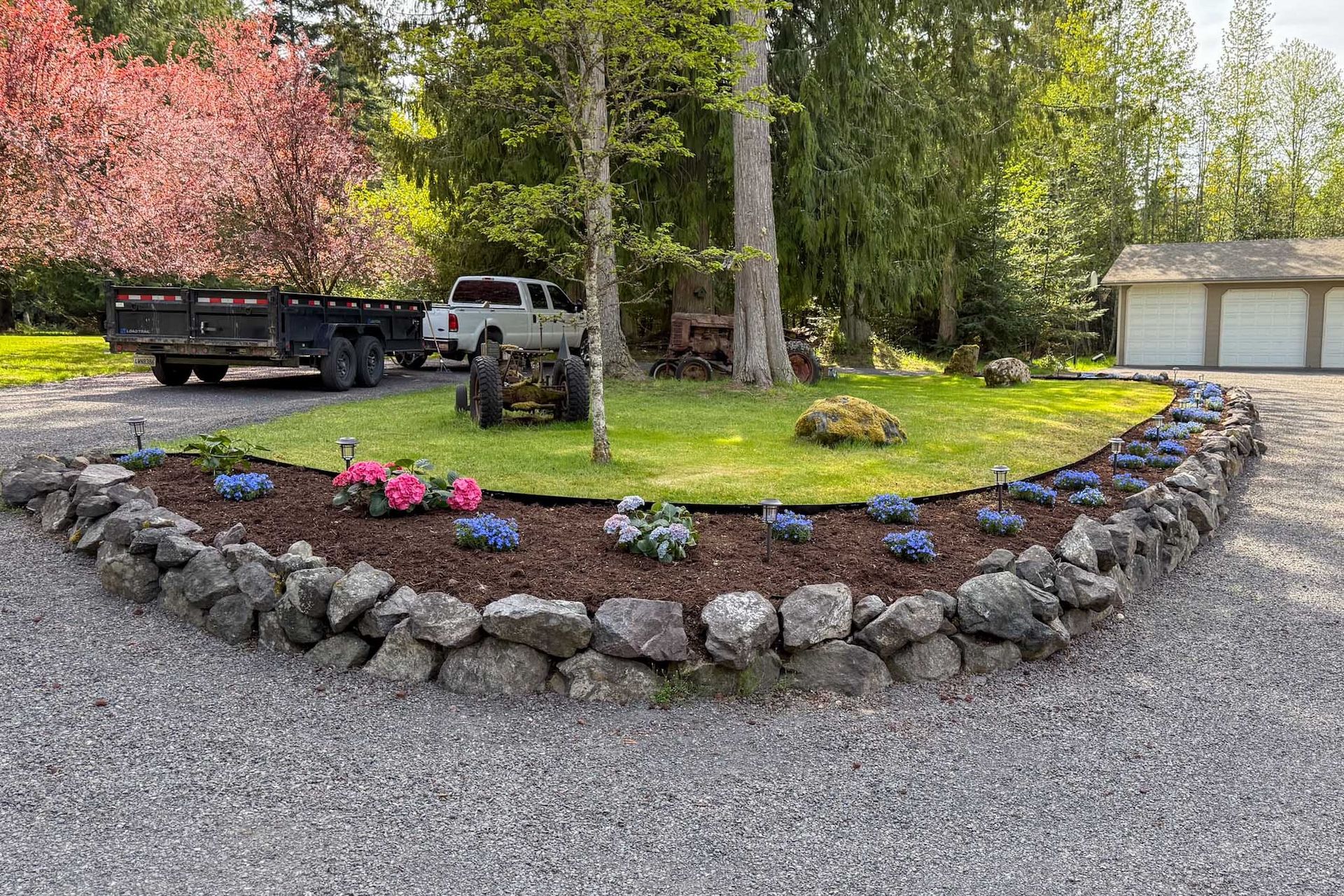A curved stone-bordered flowerbed on a gravel driveway, with a truck, trailer, and garage in the background.