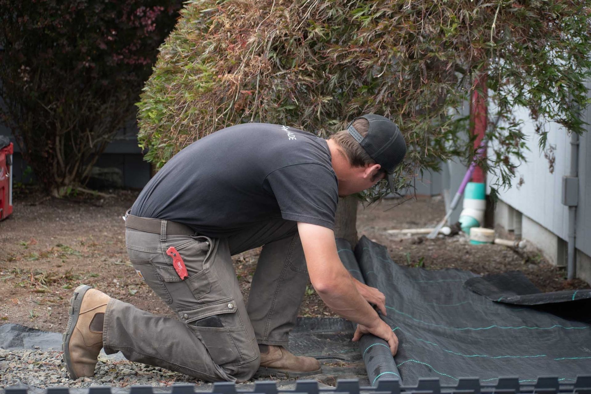 Man kneeling, installing black landscaping fabric next to a building.