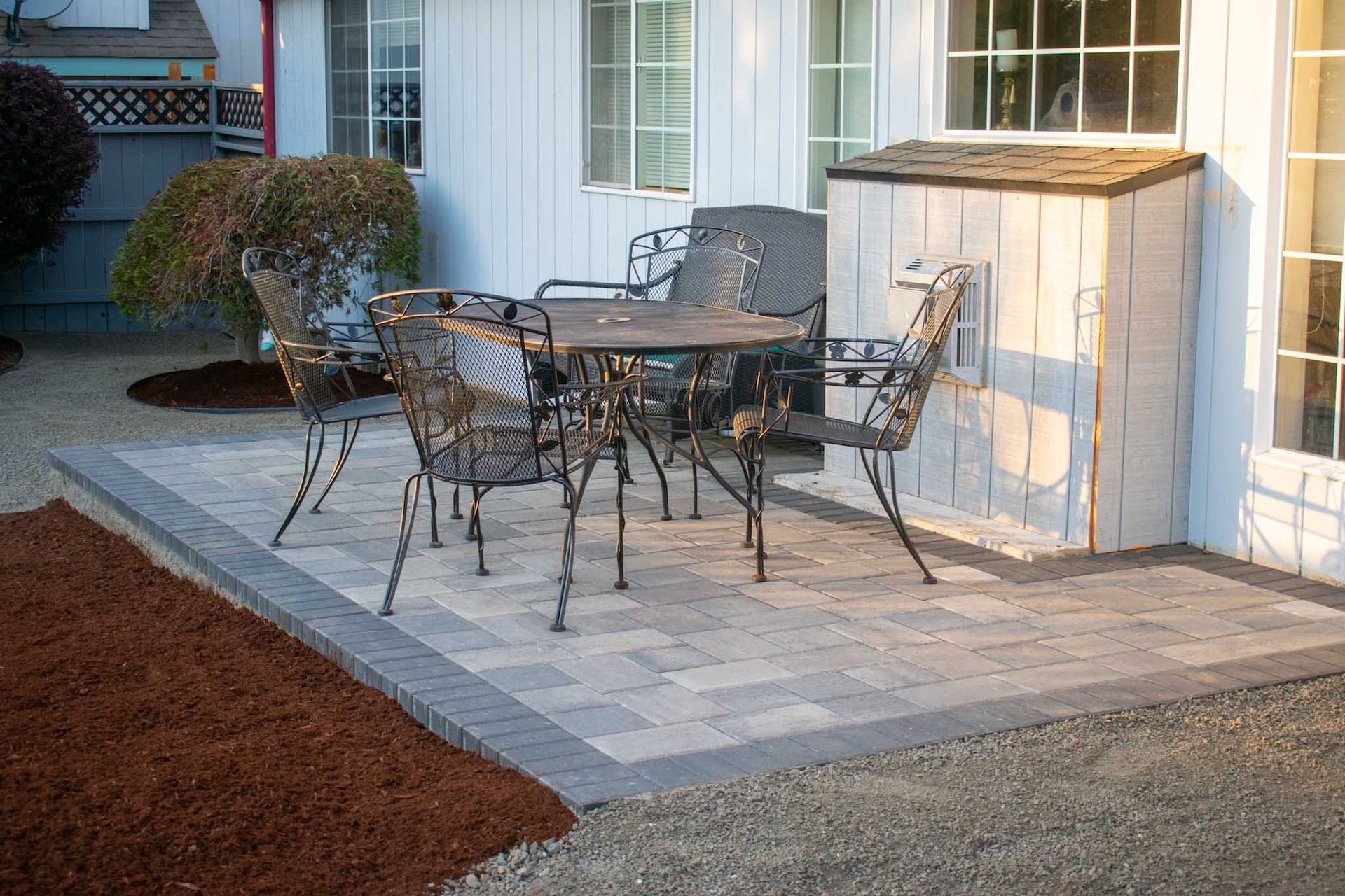 Outdoor patio with wrought iron table and chairs on a brick surface, next to a white house.