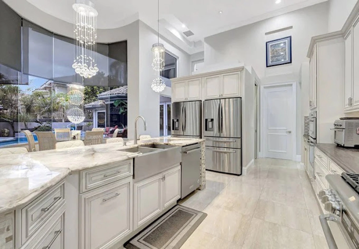 Spacious white kitchen with stainless steel appliances, crystal chandeliers, and a large window overlooking a pool.