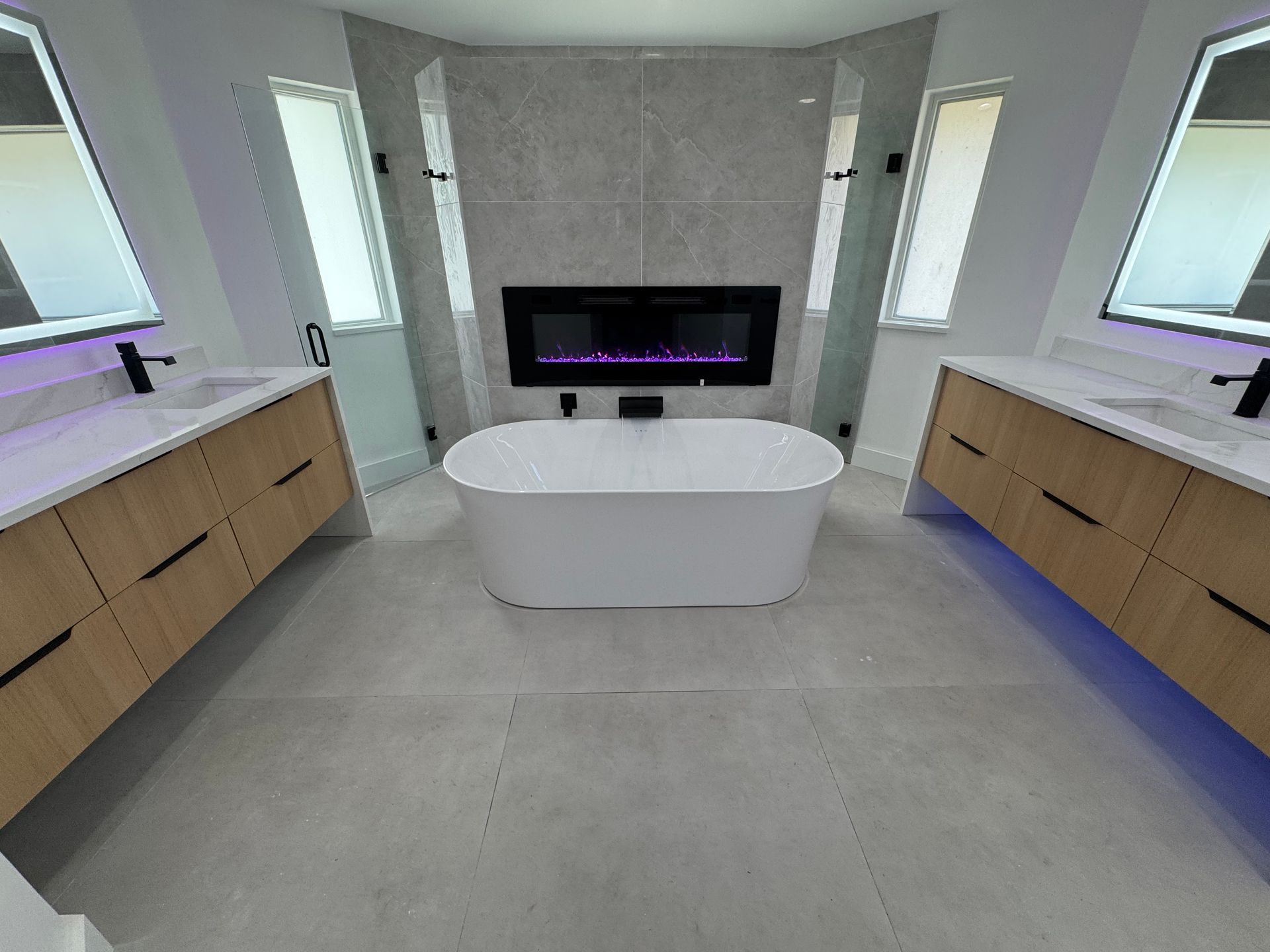 A modern bathroom with dual wood vanities, gray tile flooring, a white freestanding tub, and a built-in wall fireplace.