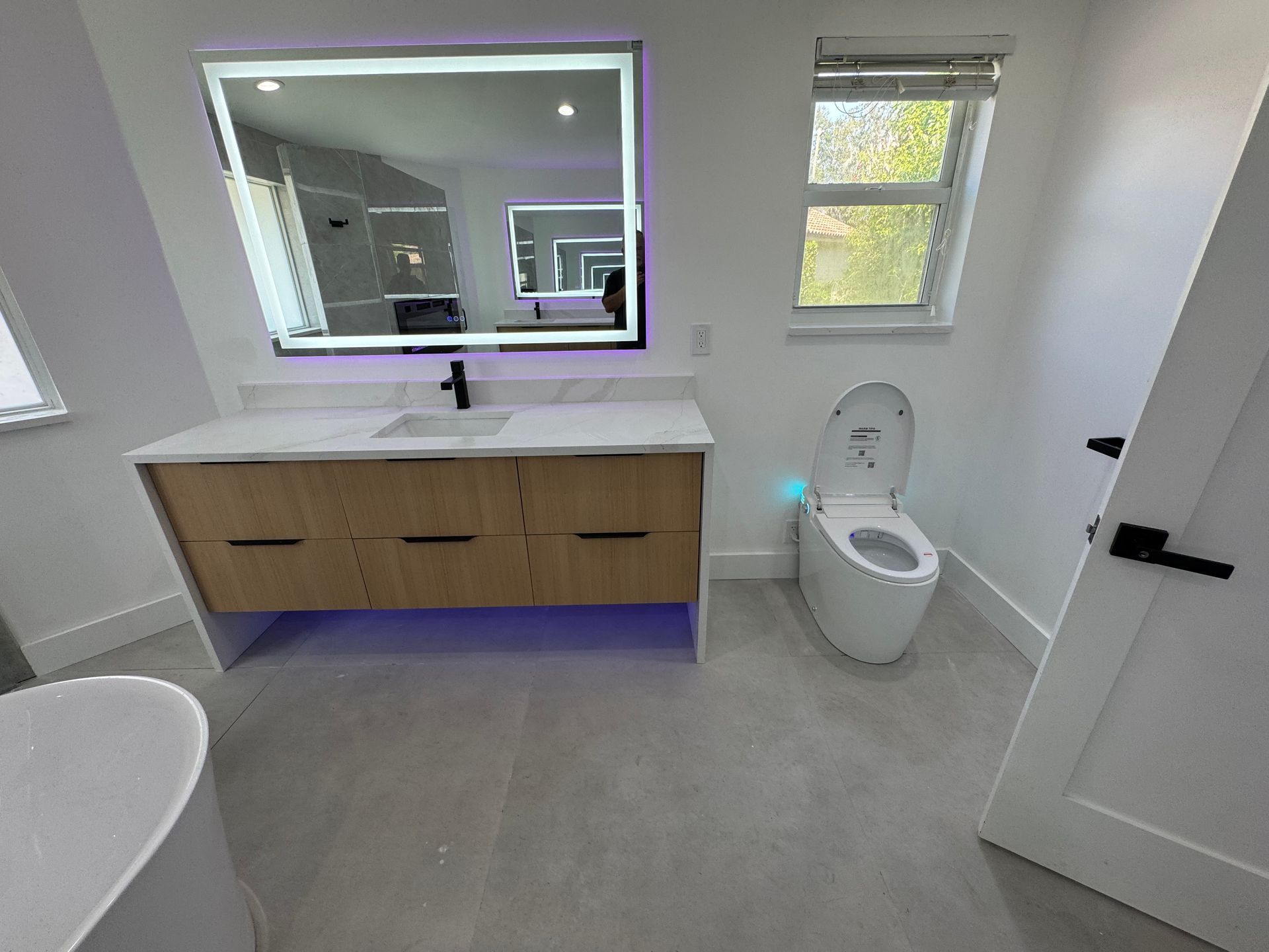 Modern bathroom with a wooden vanity, backlit mirror, a white toilet, and a bathtub in a bright, neutral-toned room.