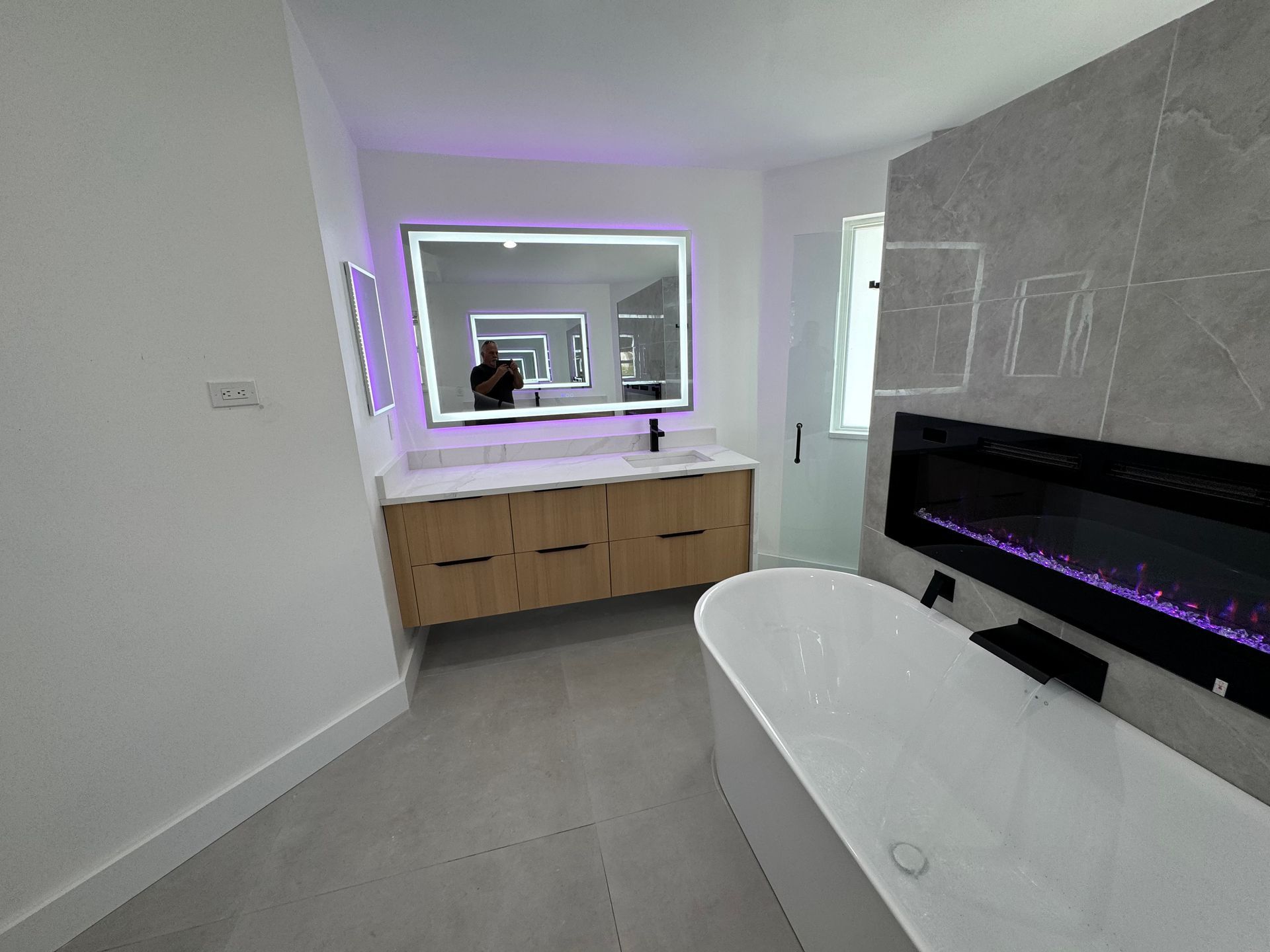 A modern bathroom with a freestanding white tub, a wall-mounted electric fireplace, and a wood vanity with LED mirror.