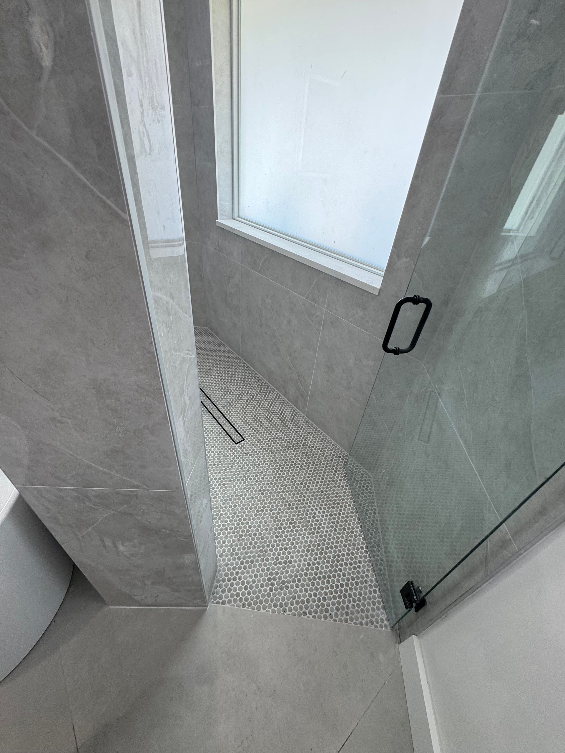 A modern shower area with large gray wall tiles, a textured pebble floor, and a glass door with a black handle.