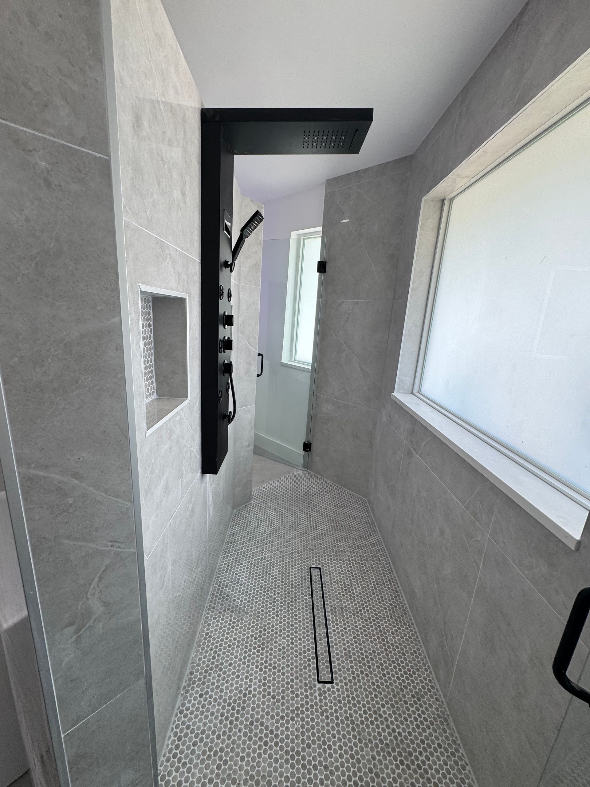 A modern walk-in shower with grey stone tiles, a black shower panel, a linear drain, and a window on the right wall.