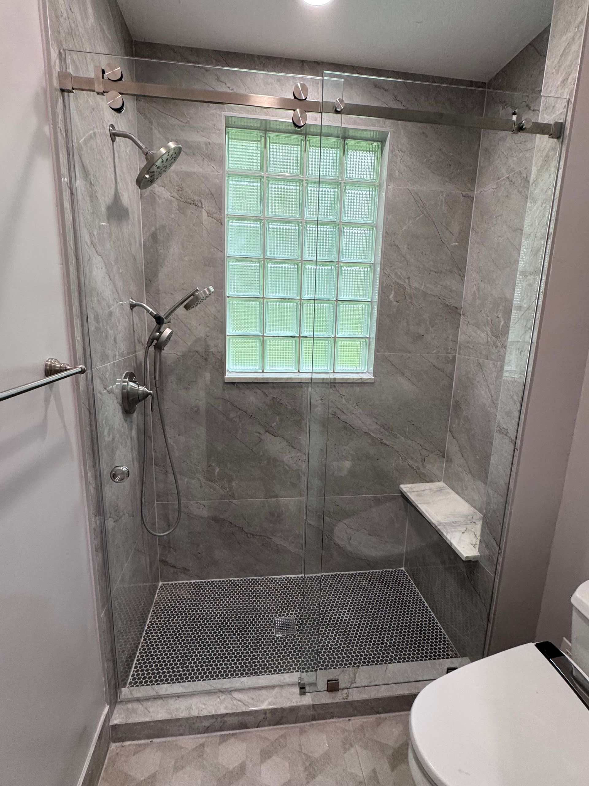 A modern walk-in shower with gray tiled walls, a glass block window, a pebble floor, a built-in bench, and sliding doors.