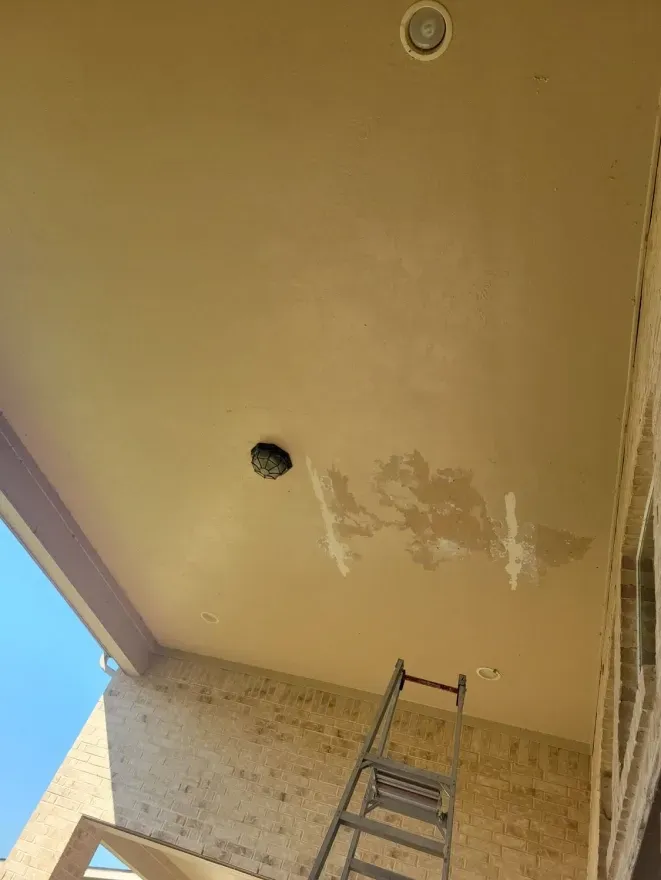 Ladder beneath a beige outdoor ceiling with peeling paint and a recessed light hole