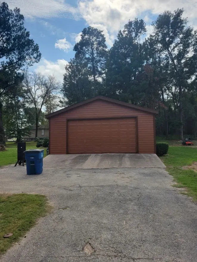 Red garage with closed door at the end of a gravel driveway, flanked by trees and a blue trash bin.