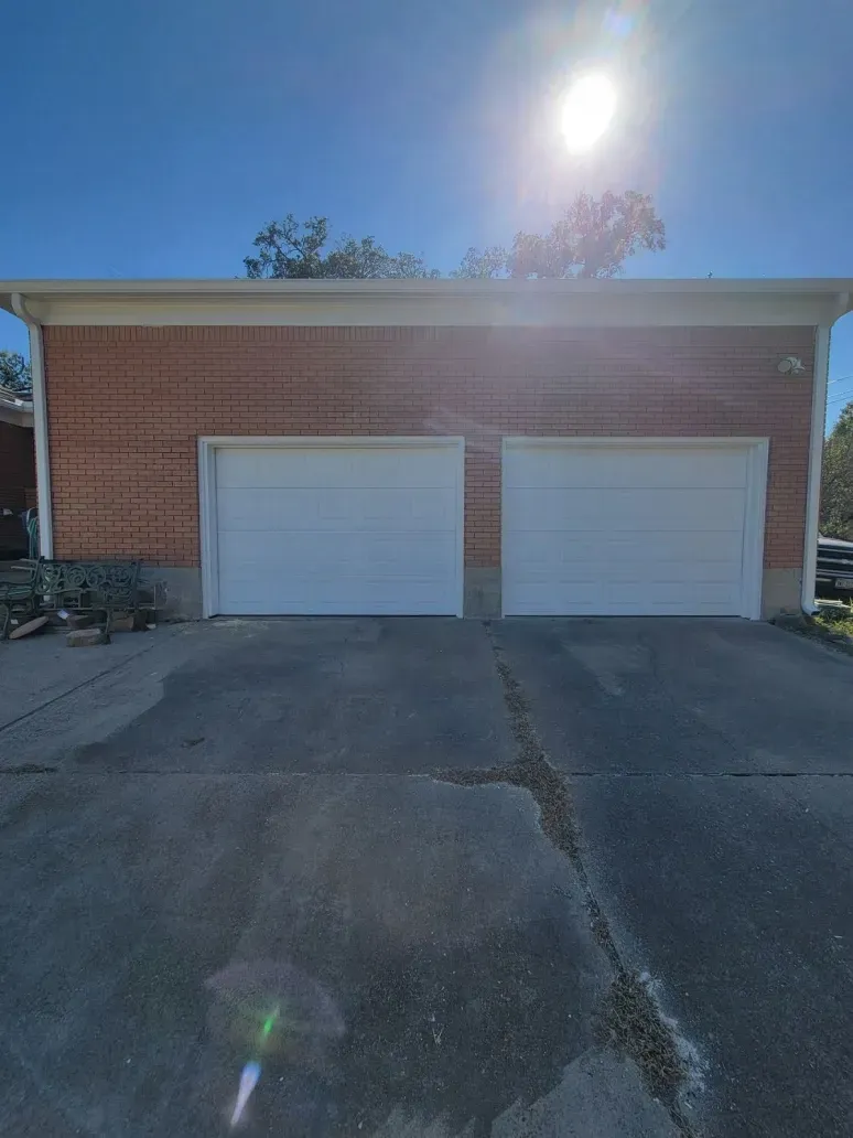 Brick garage with two white doors, seen from a driveway in bright sunlight.