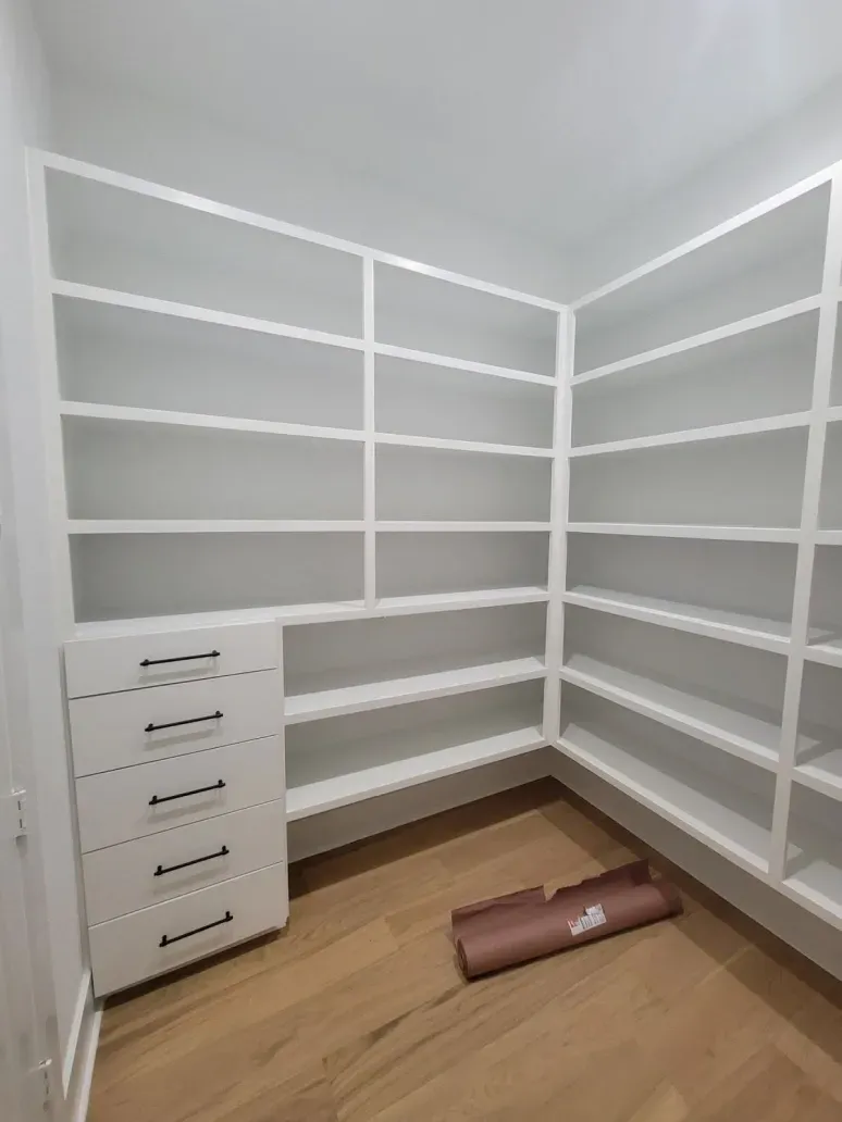 Empty white walk-in closet with built-in shelves and a small drawer unit on wood flooring.