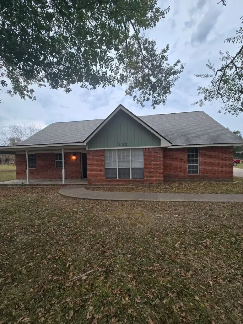 Single-story red brick house with a green gable and a large front yard under an overcast sky