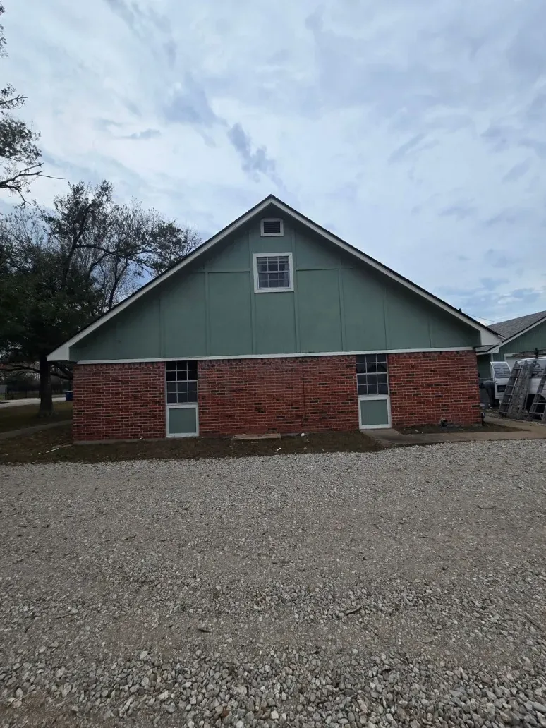 Small green house with red brick lower walls and a gravel lot in front under a cloudy sky