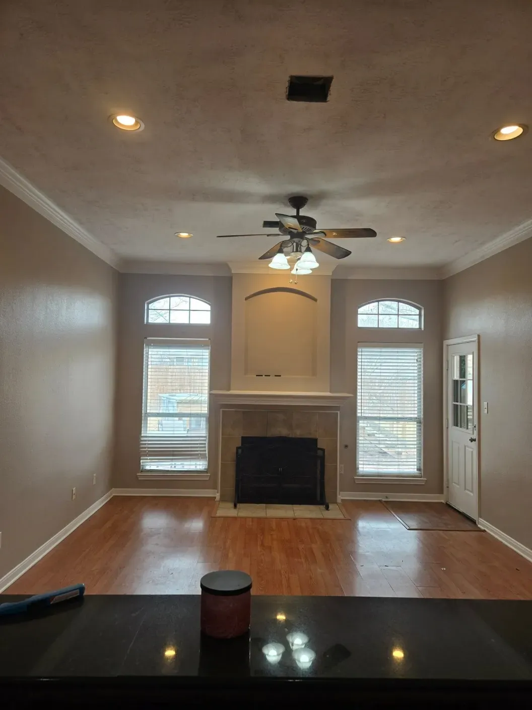 Empty living room with fireplace, arched windows, ceiling fan, and hardwood floors