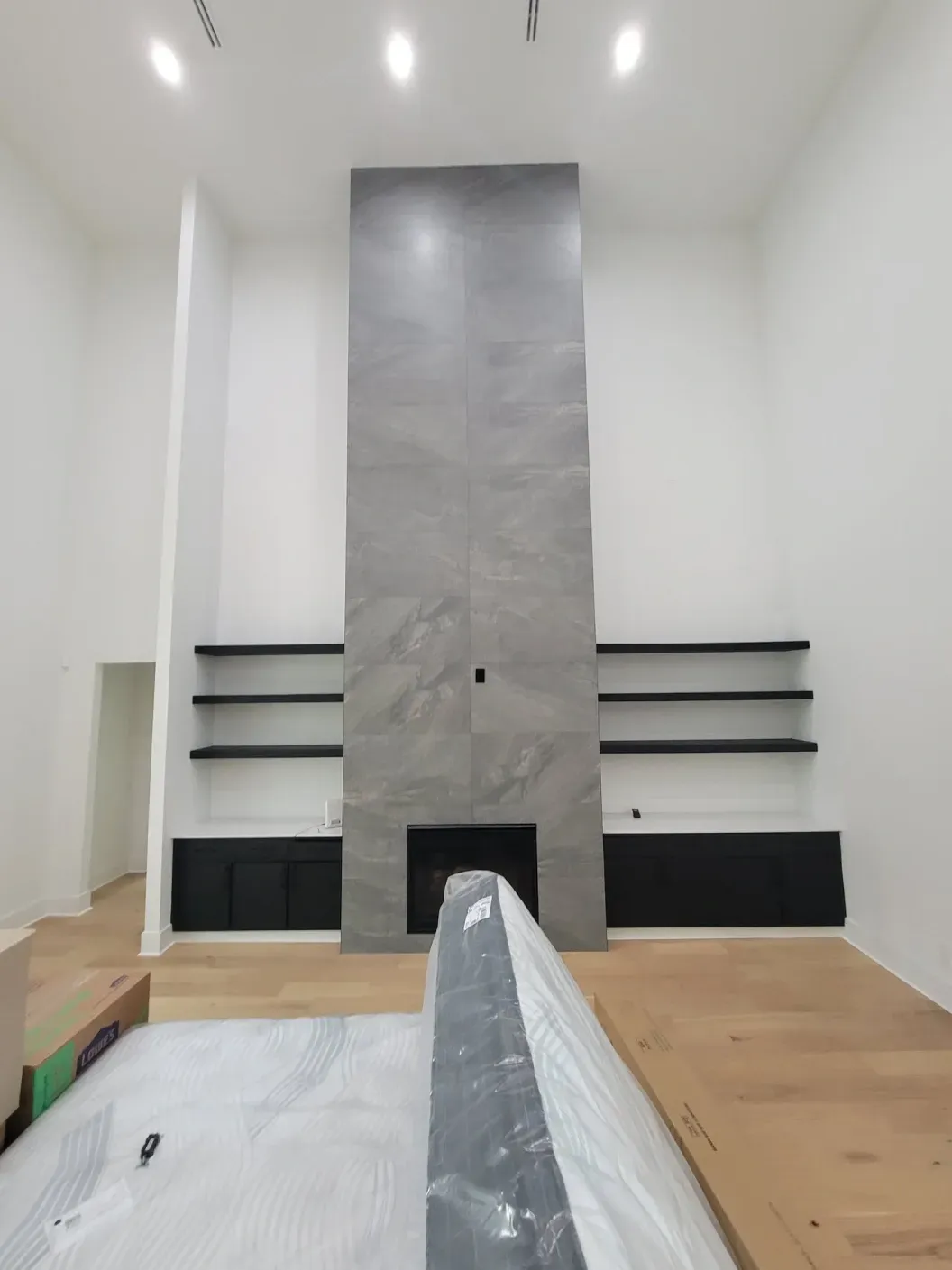 Modern living room under renovation with a tall gray fireplace, white walls, and unfinished wood floor.