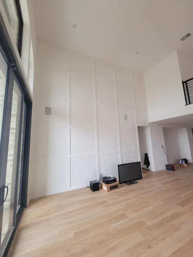 Empty bright living room with tall windows, white walls, and light wood floors; small TV and boxes on the floor.