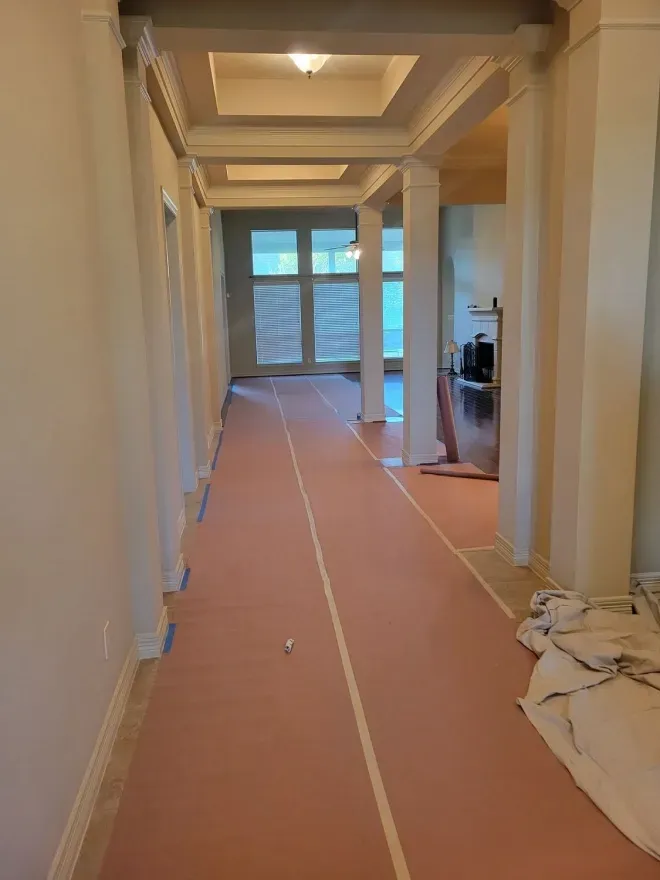 Hallway under renovation with covered floor, taped seams, and a window at the far end.