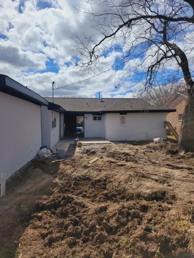 Backyard with bare dirt, white house walls, and a cloudy sky over leafless tree branches.